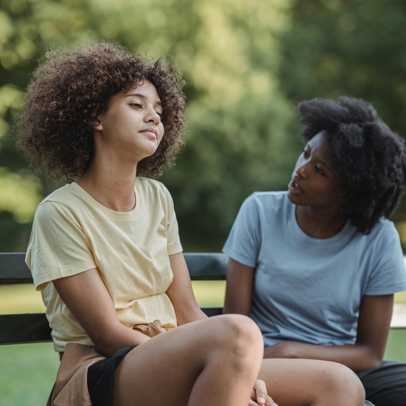 A teenage girl sits on a park bench with her arms crossed and a distant expression, while an adult woman beside her leans in, speaking with concern and empathy. The background is filled with soft, green trees, suggesting a calm outdoor setting.
